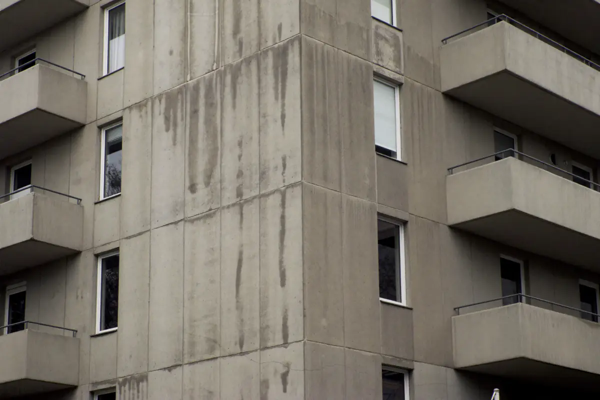 Lichtgrijze betonnen gevelpanelen van een flatgebouw uit de jaren zeventig met regensporen en haarscheuren